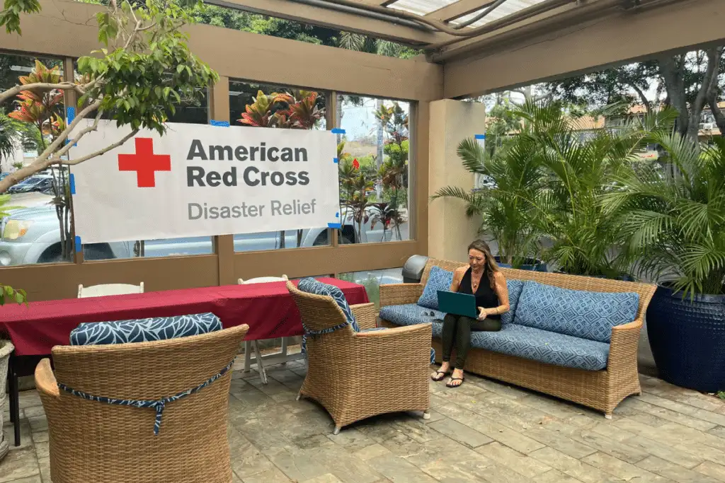 A woman sitting on a wicker patio couch, working on a laptop outdoors with a large white banner that reads "American Red Cross Disaster Relief"