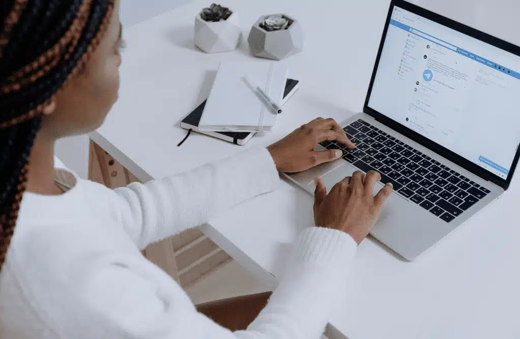 A black woman on a laptop showing a Facebook social media profile, with a notepad, a pen and two potted succulents beside her