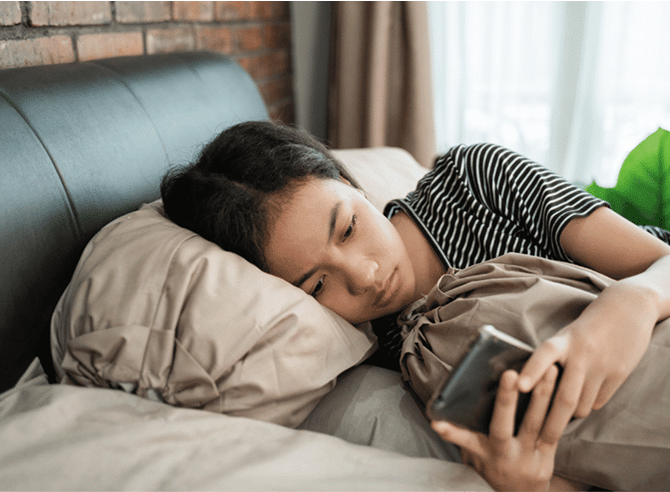 A young girl lying in bed, propped up by a pillow, looking down at her smartphone with a sad expression