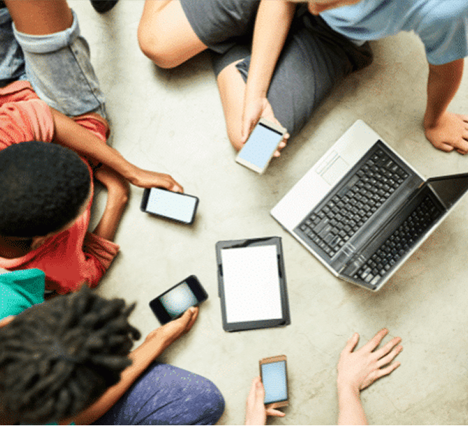An overhead shot of four people sitting on the floor, each person holding a smartphone, with a laptop and a tablet in the center