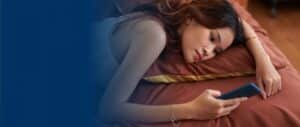 A woman lying on a bed and looking at her cell phone with a sad expression
