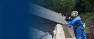 Two people in a full-body blue hazmat suit and respirator helmet on a roof performing asbestos abatement