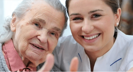 A close-up image of an elderly woman and a nurse smiling at the camera