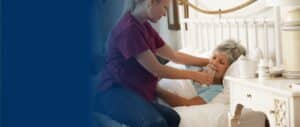 A caregiver sitting on the edge of a bed, helping a frail-looking elderly woman to drink a glass of water