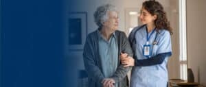 A nurse walking alongside an elderly woman, gently holding her arm and smiling at each other