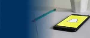 A smartphone lying on a desk with the Snapchat app icon displayed on the screen, a pencil and a white cup in the background
