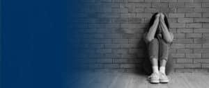 A young person sitting on a floor with their back against a brick wall, hugging their knees with their head down